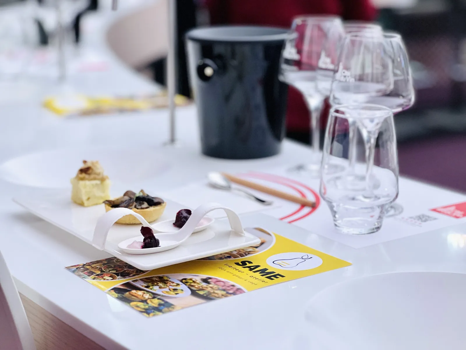 Une mise en place élégante avec une assiette blanche contenant une bouchée garnie, présentée à côté de verres à vin, une carafe noire, et une brochure jaune 'SAME STILL A MEAL' dans un cadre gastronomique.