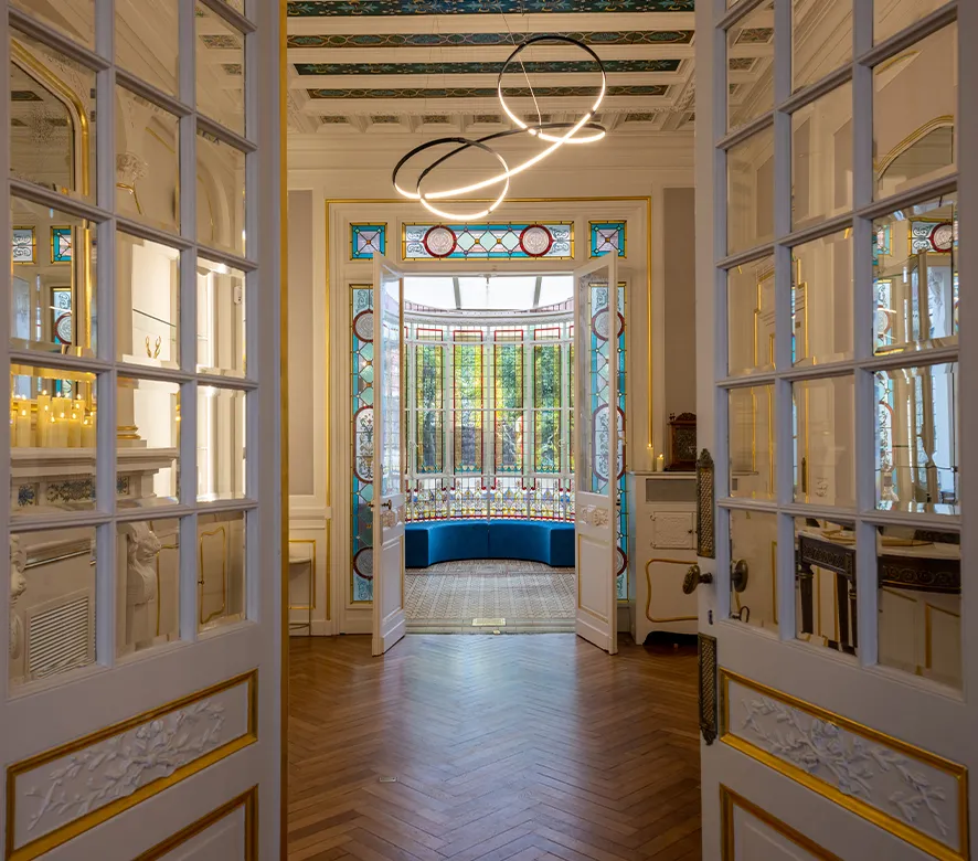 Couloir élégant avec doubles portes vitrées ouvrant sur une baie vitrée lumineuse avec assise en velours bleu, fenêtres en vitrail ouvragé, dorures et luminaires circulaires modernes, mettant en avant l'architecture raffinée de Bordeaux.