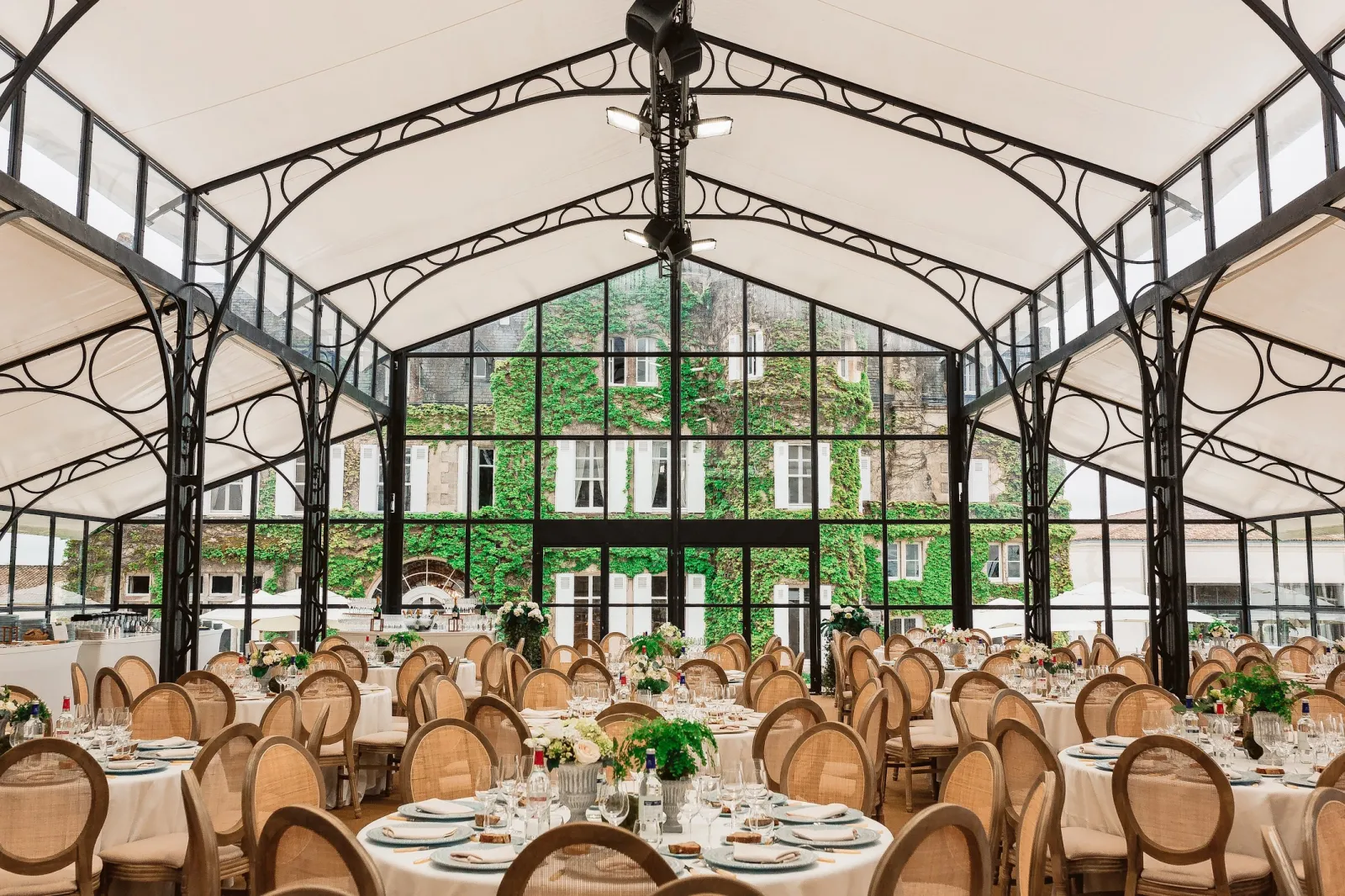 Salle de réception élégante avec verrière et structure en fer forgé, tables rondes dressées pour un événement formel, chaises en rotin, nappes blanches, et bâtiment couvert de lierre visible à travers les grandes baies vitrées à Bordeaux.