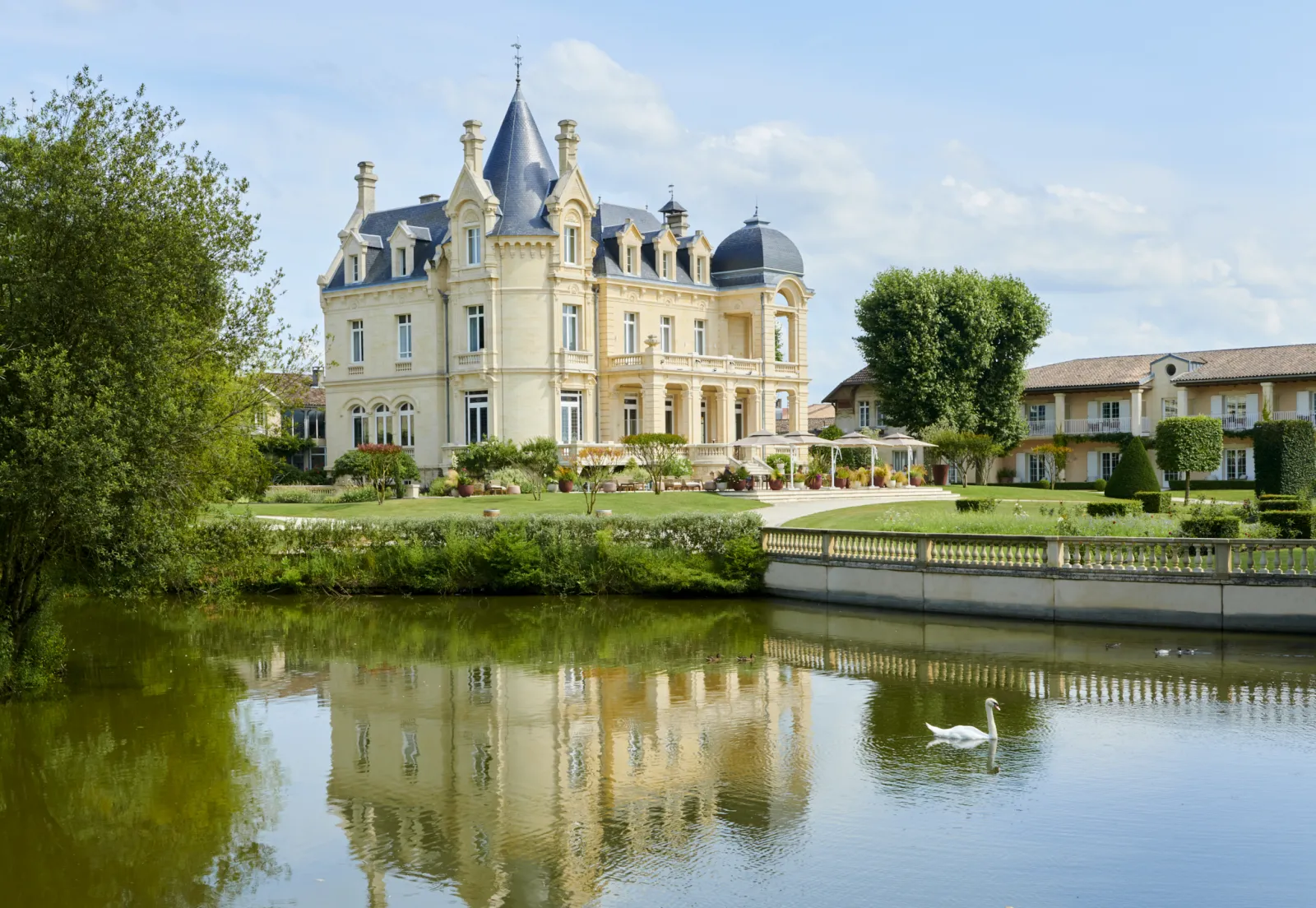 Château Hôtel Grand Barrail à Bordeaux, un manoir de style Second Empire de couleur crème aux toits mansardés bleu-gris et tourelles, se reflétant dans un étang tranquille avec jardins à la française et bâtiments adjacents