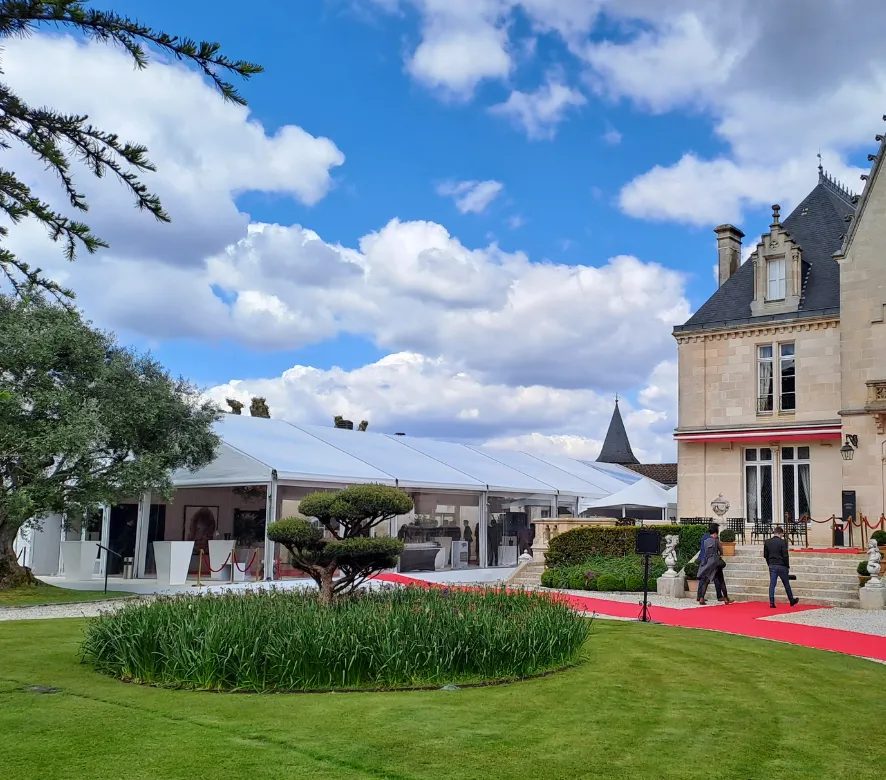Installation événementielle élégante devant un château bordelais avec tapis rouge, chapiteau blanc, jardins entretenus et bâtiment historique en pierre sous un ciel bleu