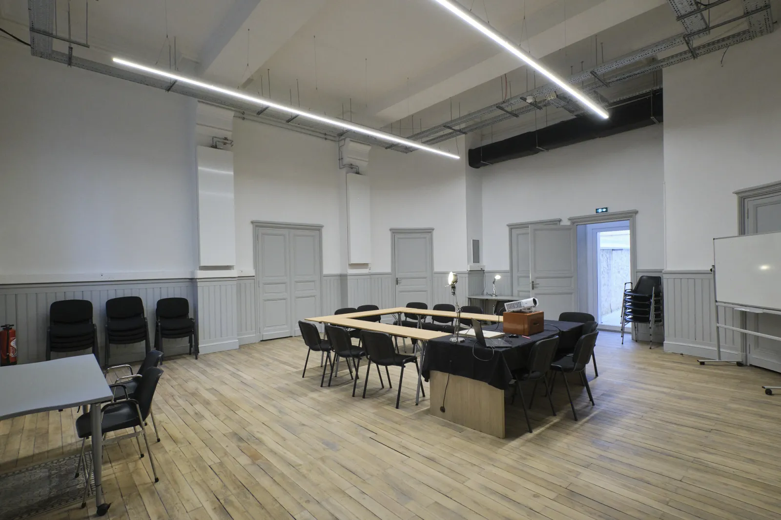Salle de réunion moderne de ManuCo Bordeaux avec parquet, grande table de conférence entourée de chaises noires, murs blancs à lambris, éclairage linéaire au plafond et plusieurs portes.