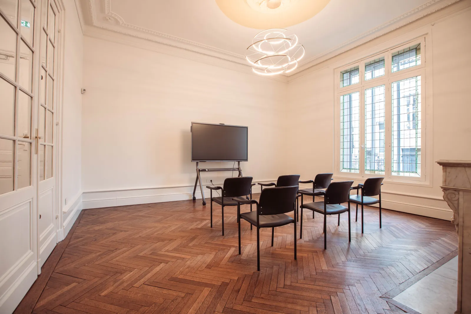 Salle de réunion moderne avec parquet en chevrons, murs blancs, luminaires contemporains, grandes fenêtres à carreaux, écran TV mural sur pied et quatre chaises noires disposées en demi-cercle.