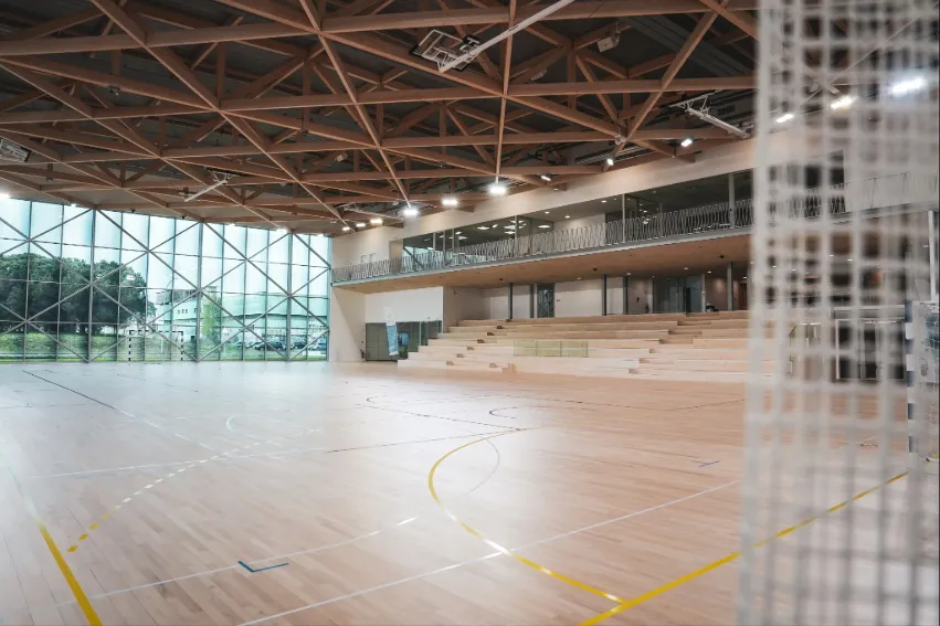 Intérieur d'un gymnase moderne de l'Université de Bordeaux avec un parquet sportif marqué, une structure de toit en acier apparente, de grandes baies vitrées, un niveau de galerie supérieure et des gradins en arrière-plan.