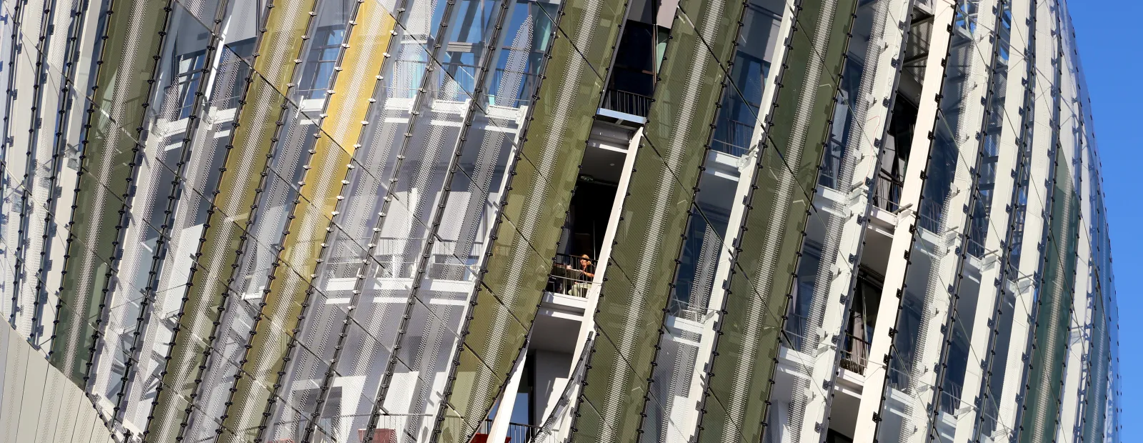 Immeuble résidentiel moderne aux formes courbes avec façade rayée verte et blanche, panneaux grillagés métalliques et balcons, photographié sous un ciel bleu dégagé à Bordeaux.