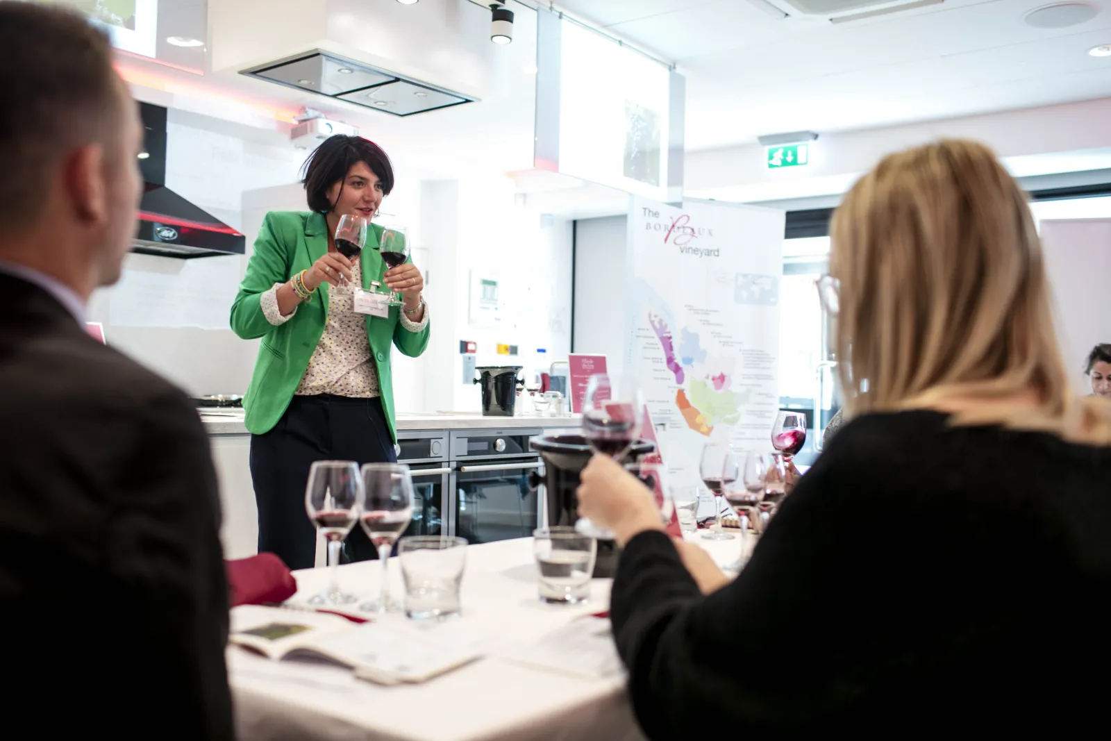 Une femme vêtue d'une veste verte vive présente devant un public dans une cuisine moderne, tenant un microphone lors d'une dégustation de vin à Bordeaux. Les participants sont assis à une table avec des verres de vin et du matériel pédagogique sur les rég