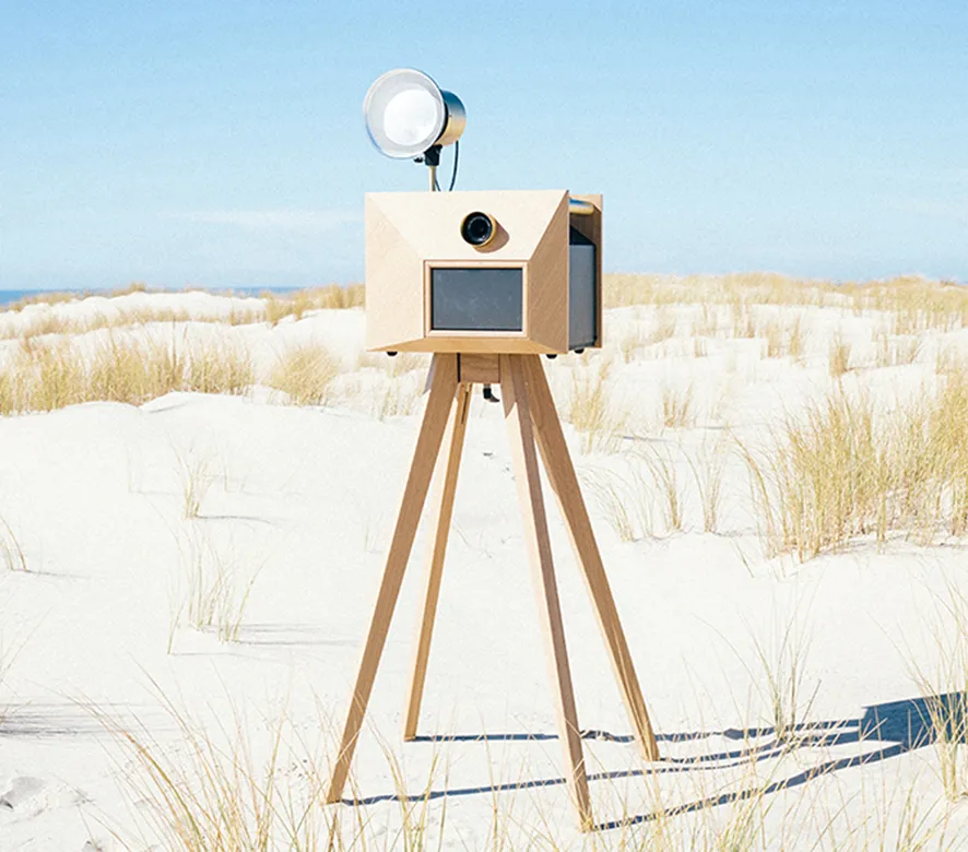 Installation d'un appareil photo cabine en bois sur trépied dans un paysage de plage avec dunes et oyats sous un ciel bleu dégagé