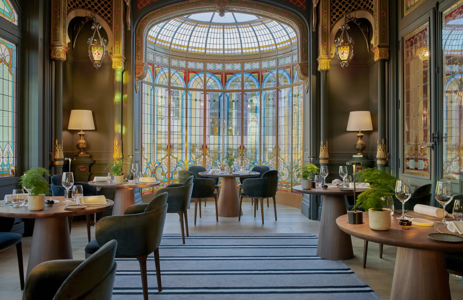 Salle de restaurant élégante avec dôme en vitrail Art Déco, tables rondes en bois avec chaises noires, appliques murales en laiton ouvragé, et plantes décoratives dans un établissement hôtelier de luxe à Bordeaux.