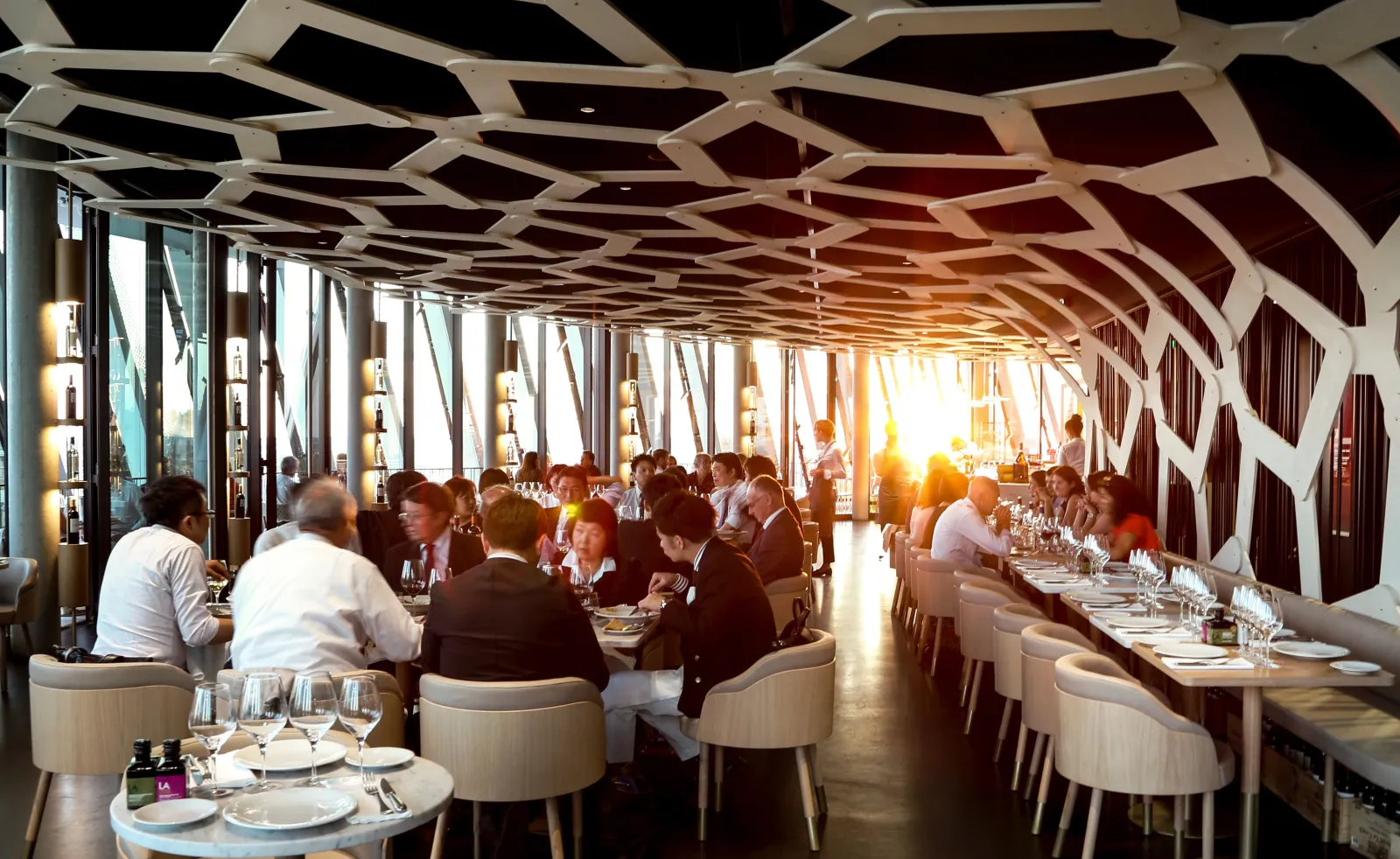 Salle de restaurant panoramique du 7 à La Cité du Vin à Bordeaux, avec des convives assis aux tables sous un plafond géométrique en bois, baies vitrées donnant sur la ville au coucher du soleil