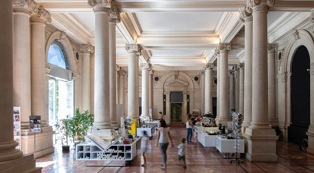 Intérieur du Musée d'Aquitaine à Bordeaux montrant un grand hall néoclassique avec de hautes colonnes corinthiennes, un plafond orné, et des visiteurs explorant des espaces d'exposition contemporains avec des unités d'exposition blanches.