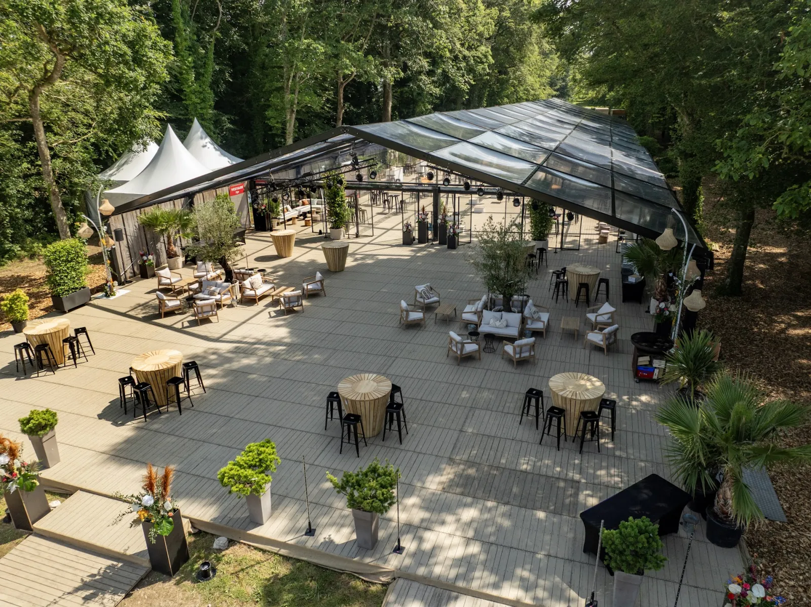 Vue aérienne d'un espace événementiel haut de gamme à Bordeaux entouré d'arbres, avec un pavillon moderne à toiture rétractable, tables de restaurant, zones de détente et jardinières décoratives sur une terrasse en bois.