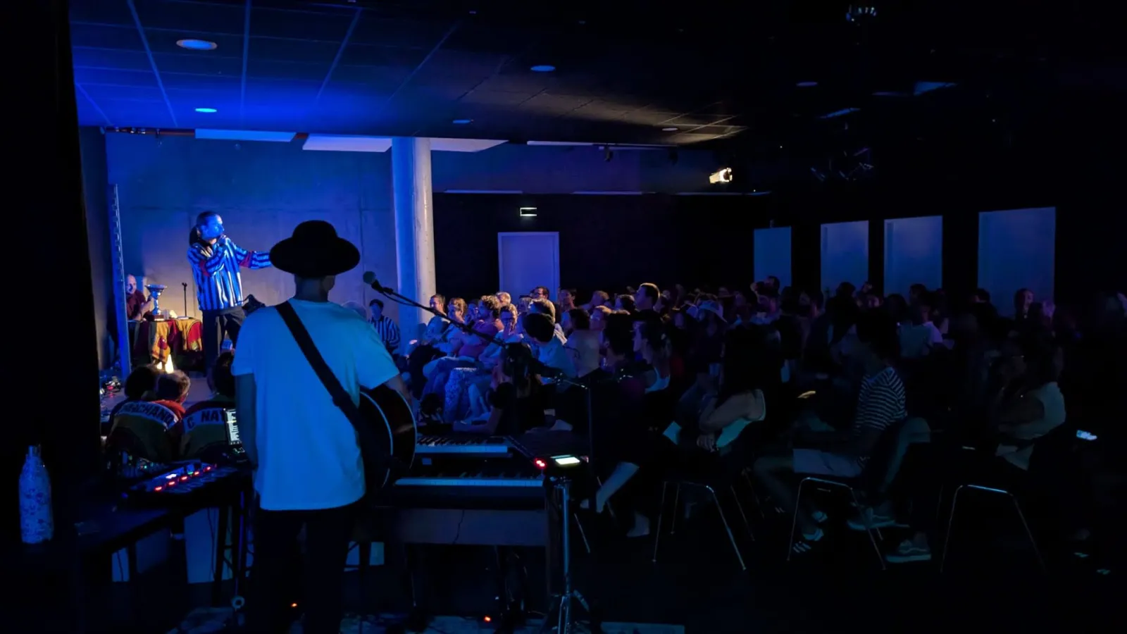 Concert en intérieur à L'Estrade à Bordeaux avec un musicien portant un chapeau de cowboy jouant de la guitare sur scène devant un public attentif dans une salle éclairée en bleu