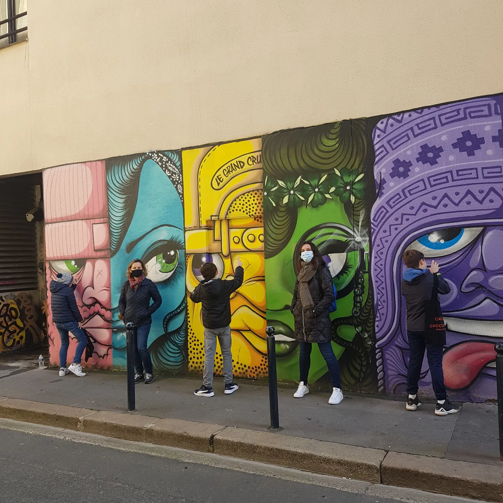 Cinq personnes posées devant une fresque murale colorée représentant des visages stylisés en rose, turquoise, jaune, vert et violet sur une façade à Bordeaux.