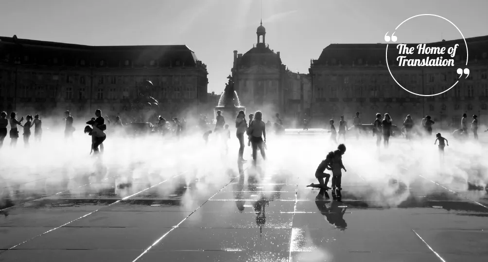 Photographie en noir et blanc d'une place publique à Bordeaux envahie par le brouillard des fontaines d'eau, avec des silhouettes de personnes marchant et jouant dans les jets, des immeubles historiques et une église à dôme en arrière-plan.