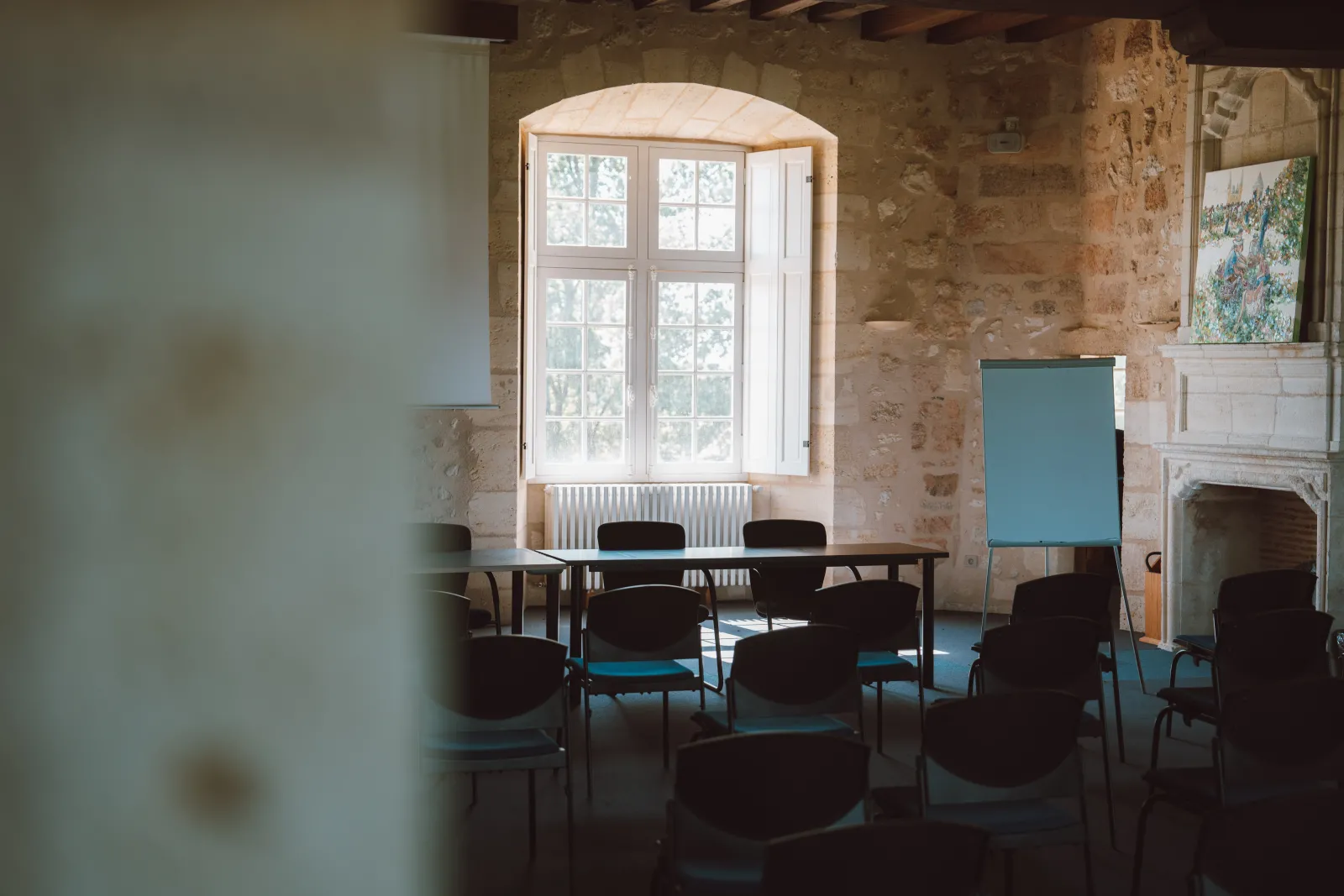 Une salle de réunion du Château d'Agassac à Bordeaux avec une table en bois, des chaises noires, de grandes fenêtres cintrées donnant sur le parc, des murs en pierre apparente, une cheminée et du mobilier bleu moderne.