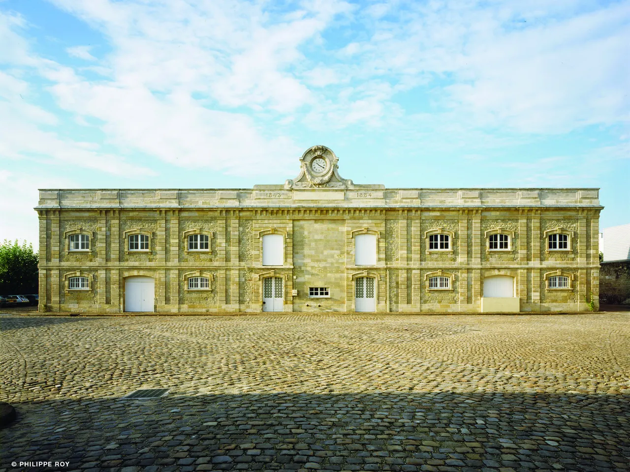 Bâtiment historique en pierre avec façade symétrique, horloge au centre, fenêtres cintrées et portes blanches, cour pavée en premier plan, Bordeaux