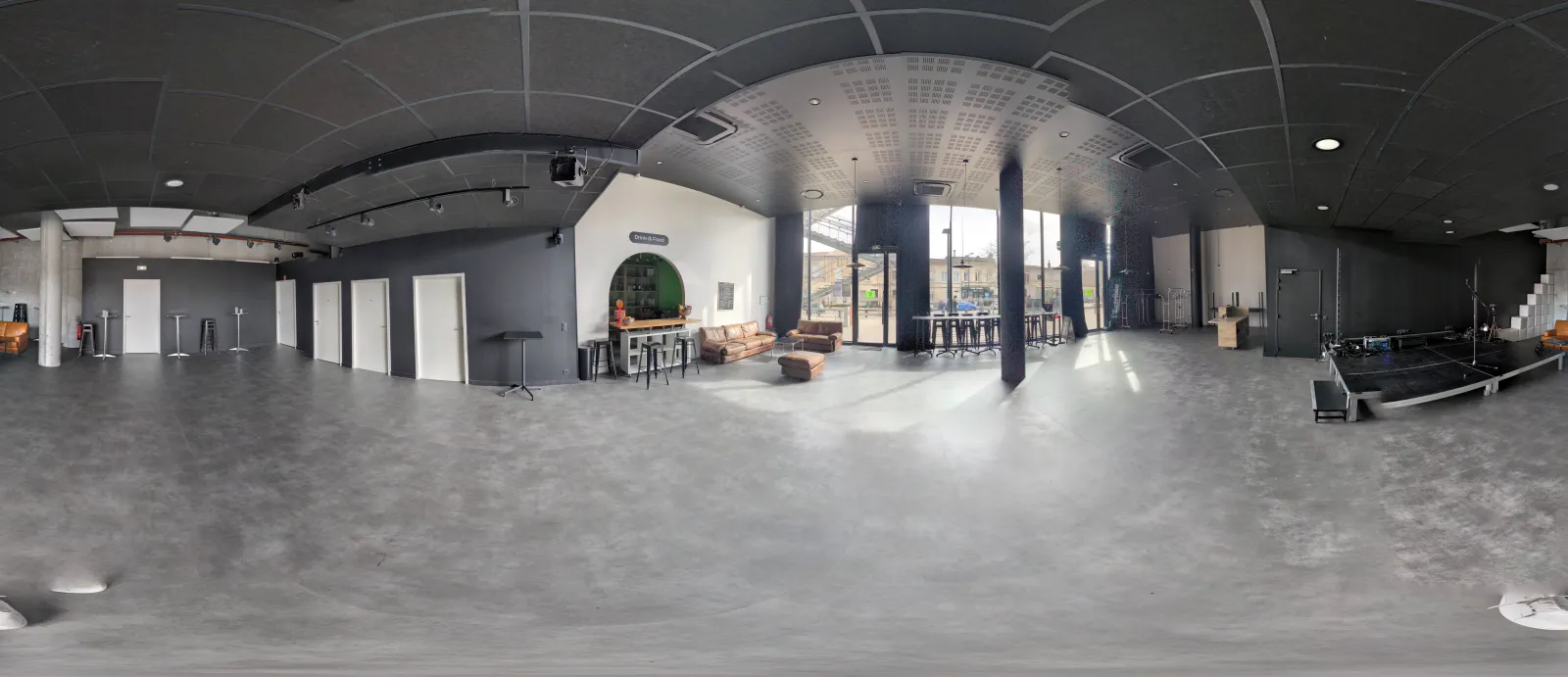 Vue panoramique d'un loft industriel moderne avec sol en béton, colonnes acier noir apparentes, plafond voûté, cloisons blanches, zone lounge et grandes baies vitrées donnant sur l'extérieur.