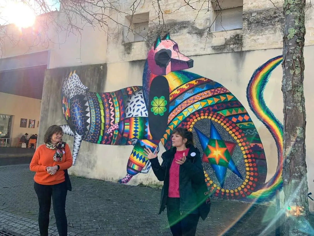 Deux femmes admirant une fresque murale représentant un oiseau stylisé aux motifs géométriques colorés et détails arc-en-ciel sur un bâtiment de Bordeaux