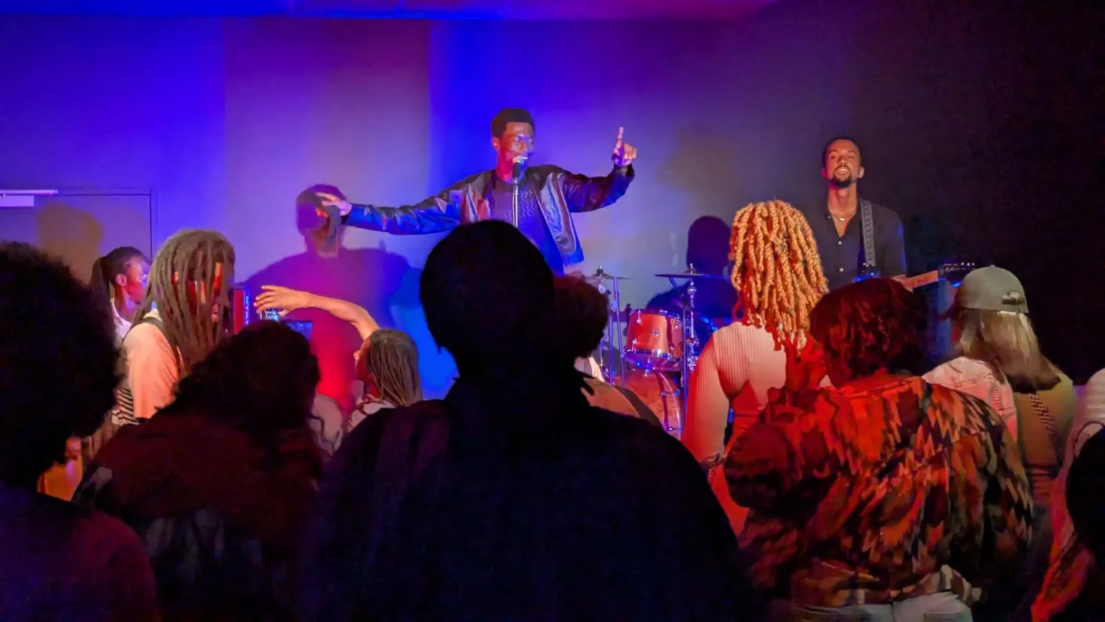 Spectacle musical en direct à L'Estrade à Bordeaux, avec un chanteur en veste métallisée sur scène, un groupe avec batterie en arrière-plan, et un public enthousiaste au premier plan sous des lumières de scène bleues et rouges.
