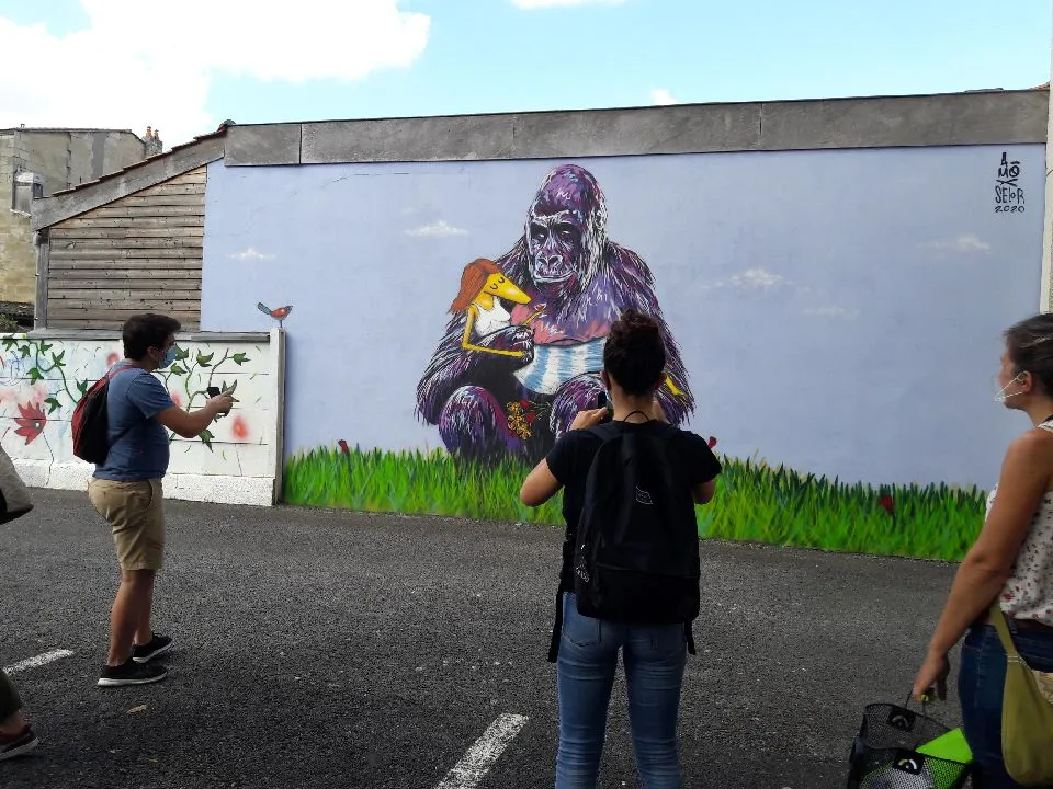 Fresque murale à Bordeaux représentant un grand gorille violet tenant un perroquet coloré, entouré d'herbe verte, avec des visiteurs photographiant l'œuvre