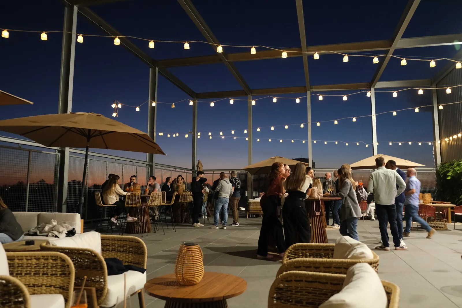 Événement en soirée sur la terrasse d'EVA Bordeaux Bègles avec des invités discutant sous des guirlandes lumineuses et des structures de pergola, mobilier en rotin au premier plan, panorama urbain visible au crépuscule