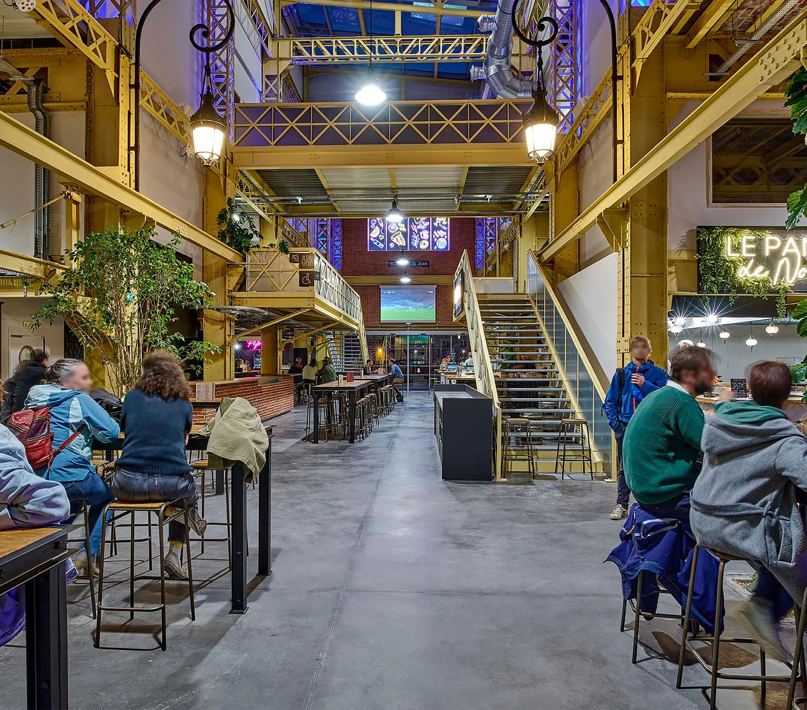 Intérieur d'une halle alimentaire moderne aux structures jaunes industrielles, zones de restauration avec clients assis aux tables, escalier, plantes en pots et éclairage ambiant nocturne.