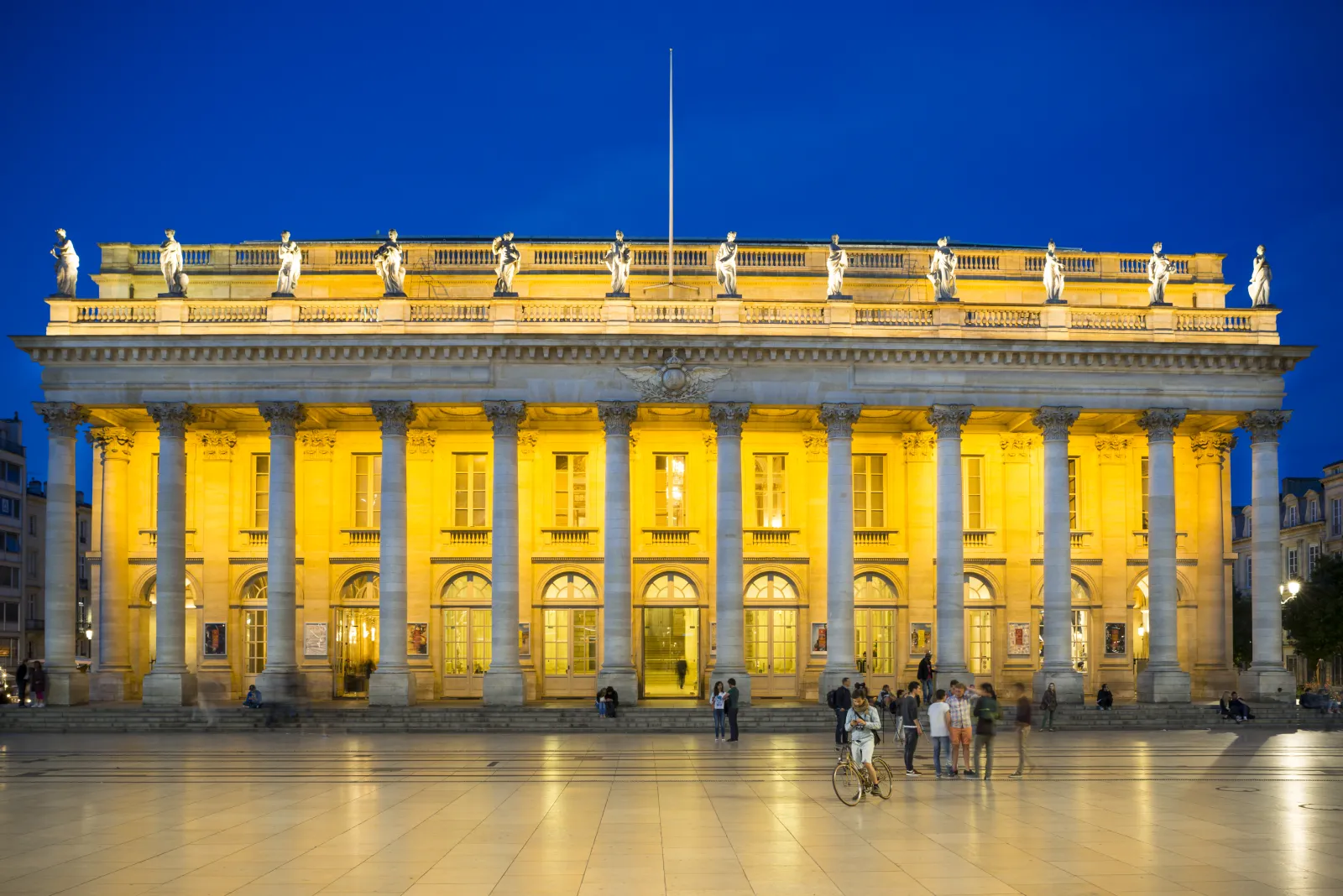 Le Grand Théâtre de Bordeaux illuminé la nuit, présentant une architecture classique avec des colonnes corinthiennes, une façade dorée, des statues sur la toiture et des visiteurs sur la place sous un ciel bleu profond.