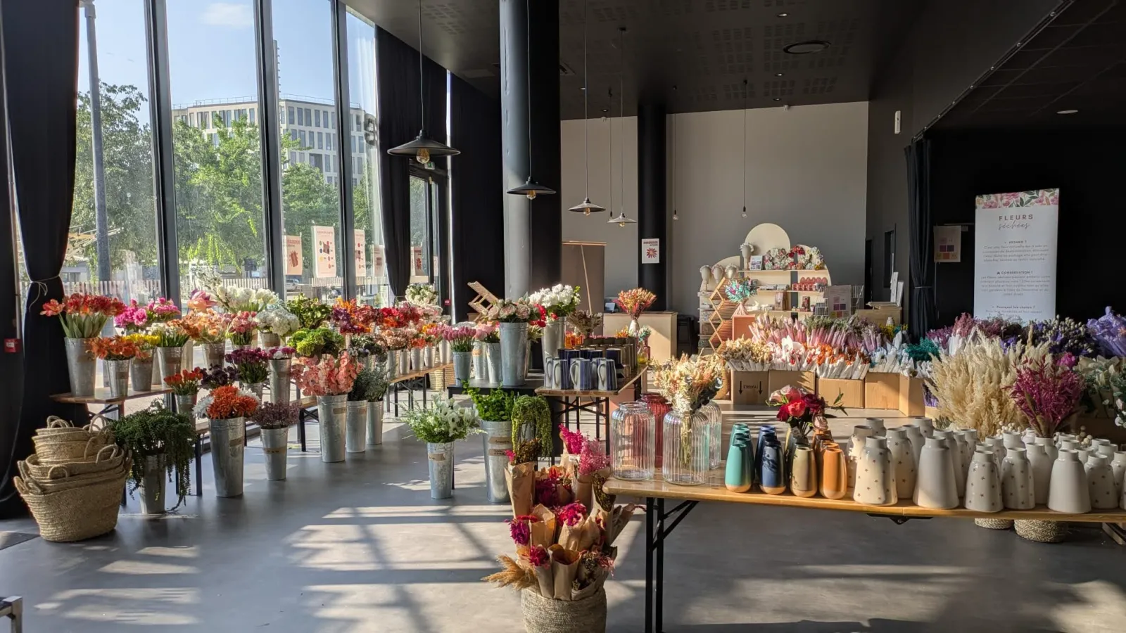 L'intérieur lumineux d'une fleuristerie moderne à L'Estrade à Bordeaux, présentant de nombreuses fleurs fraîches et séchées colorées exposées dans des vases blancs et des seaux métalliques, avec des étagères en bois, des luminaires suspendus et de grandes