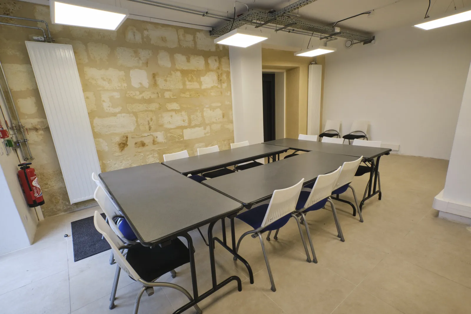 Salle de réunion contemporaine à La ManuCo à Bordeaux avec tables de conférence modulables, chaises blanches, mur en pierre apparente, panneaux LED au plafond et équipements de sécurité incendie.