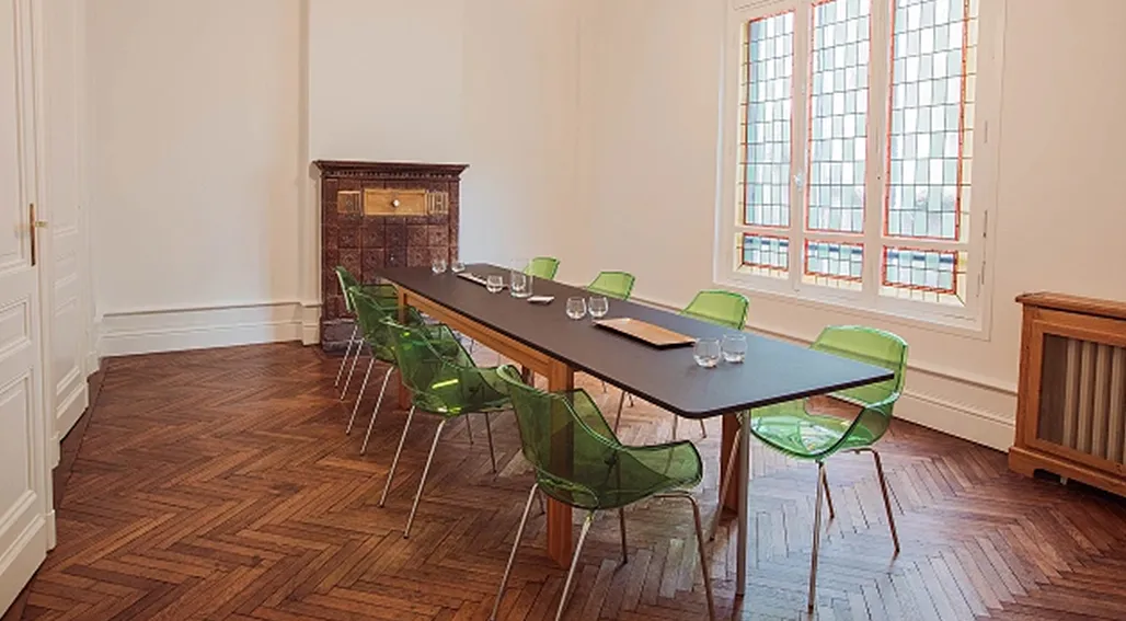 Salle de réunion moderne avec une longue table noire, des chaises transparentes vertes, un parquet en chevrons et de grandes fenêtres à carreaux dans un bâtiment historique de Bordeaux
