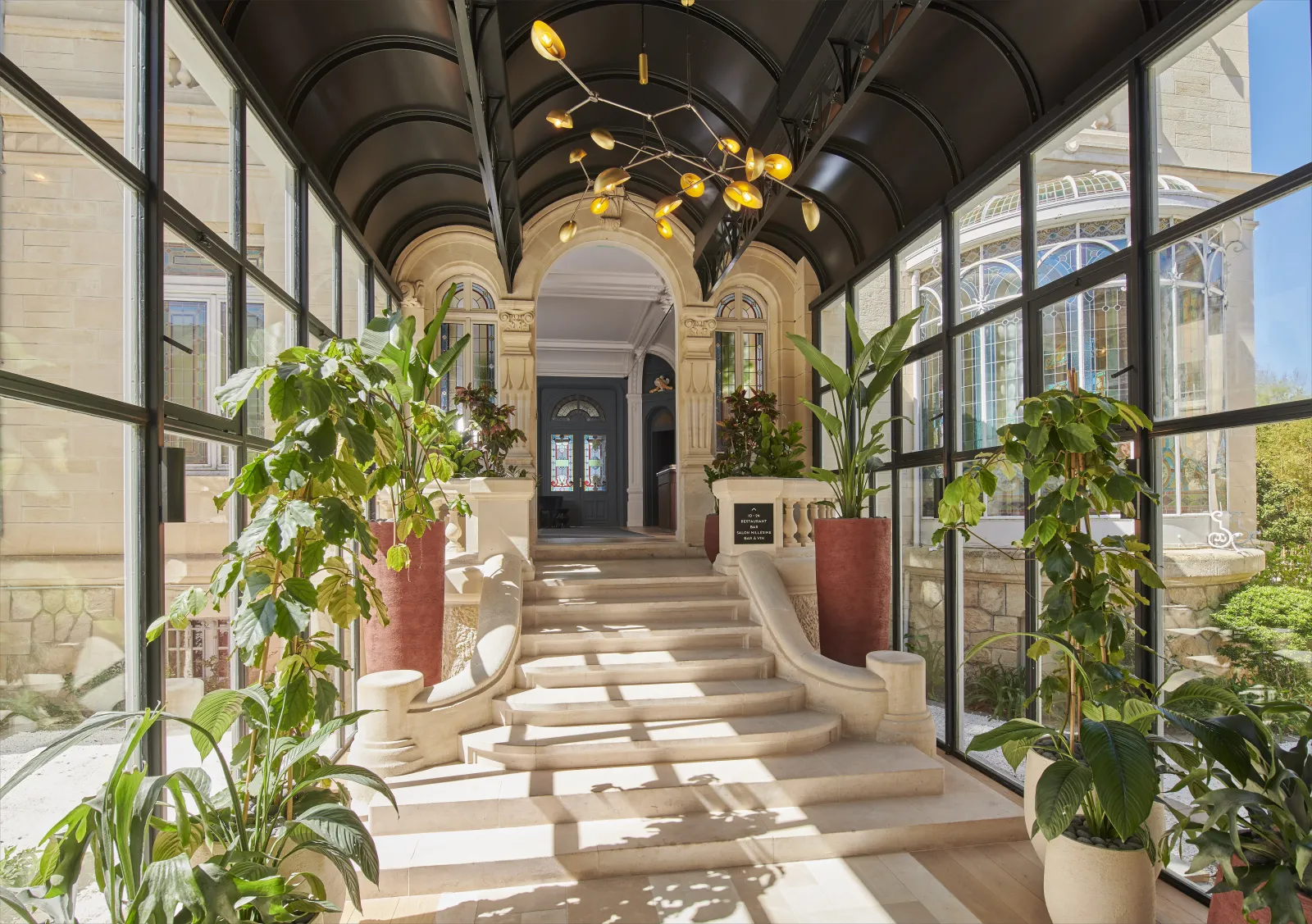 Galerie d'entrée élégante du Château Hôtel Grand Barrail à Bordeaux, avec un plafond en voûte de barrique noir, lustres dorés, grandes baies vitrées des deux côtés, sol en pierre beige avec motifs de lumière, porte cintrée au fond, et plantes décoratives 