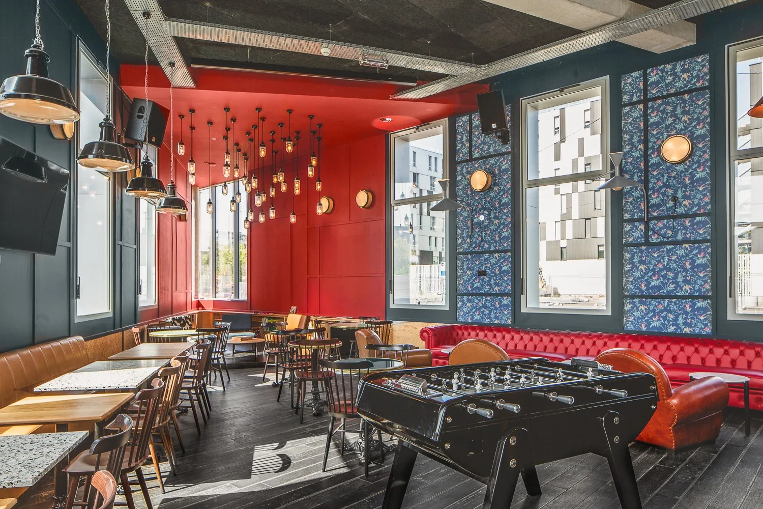 Espace lounge moderne de l'hôtel Eklo Bordeaux avec murs rouges, luminaires suspendus, baby-foot, canapé en cuir rouge, chaises en bois et grandes fenêtres sur la ville