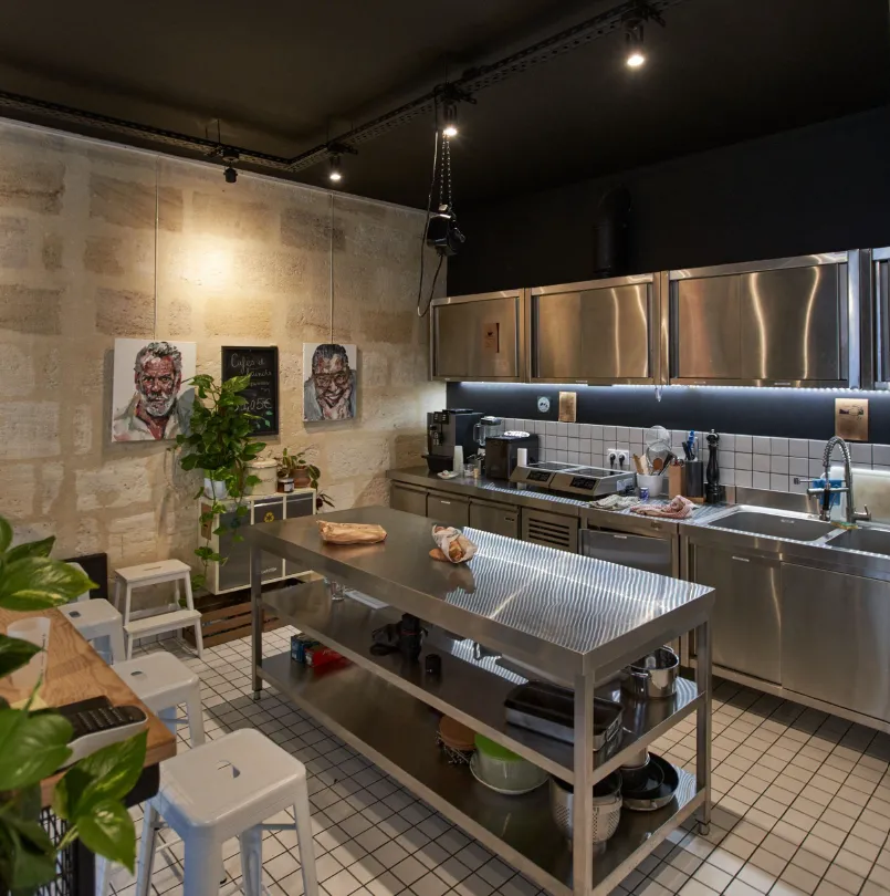 Intérieur de cuisine professionnelle avec tables de travail en acier inoxydable, équipements de cuisson, carrelage blanc, œuvres d'art sur murs beige, plantes en pot et éclairage sur rail à Bordeaux.