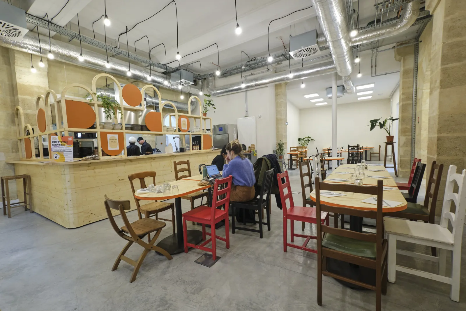 Intérieur d'un espace café contemporain à la ManuCo à Bordeaux, présentant un comptoir en bois clair avec des accents orange, des zones de sièges variées avec des chaises en bois et rouges, un sol en béton, et un plafond industriel moderne avec des lumina