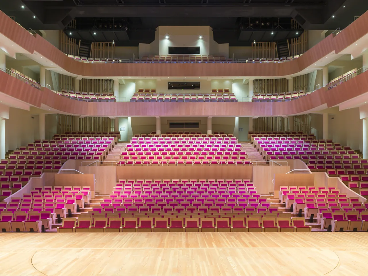 Vue intérieure du Grand Théâtre de Bordeaux montrant l'auditorium avec ses étages de sièges roses et bordeaux, la scène en bois, les balcons ornés de rampes dorées et les éléments architecturaux en crème et beige sous éclairage scénique.
