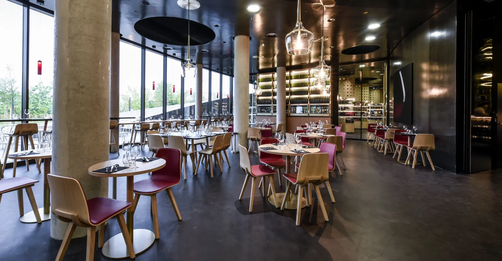 Intérieur de restaurant contemporain avec chaises en bois, assises bordeaux, tables rondes, suspensions lumineuses et bar bien garni au fond; grandes baies vitrées donnant sur la verdure.