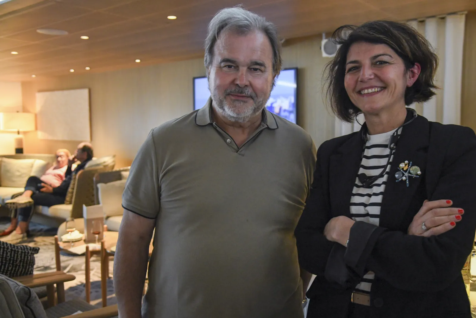 Deux personnes souriantes lors d'un événement social dans un élégant salon avec éclairage chaleureux et mobilier moderne, d'autres invités visibles en arrière-plan.