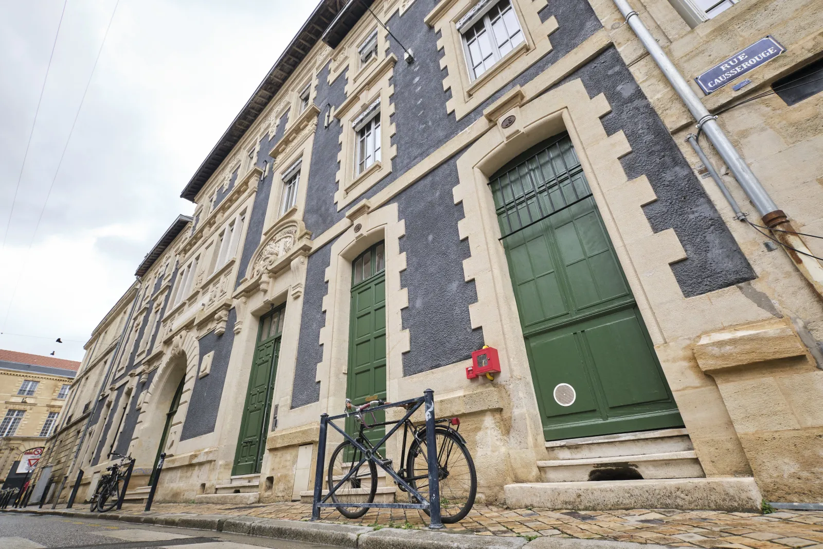 Un vélo noir garé devant un immeuble historique en pierre avec des portes vertes et des façades bleu-gris dans une rue de Bordeaux