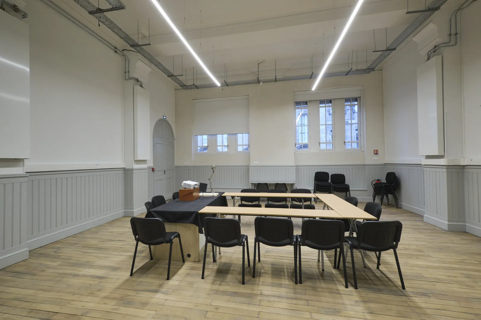 Salle de réunion contemporaine à La ManuCo à Bordeaux avec une grande table de conférence en bois, chaises noires, murs blancs à lambris, éclairage linéaire au plafond et grandes fenêtres donnant sur la ville.