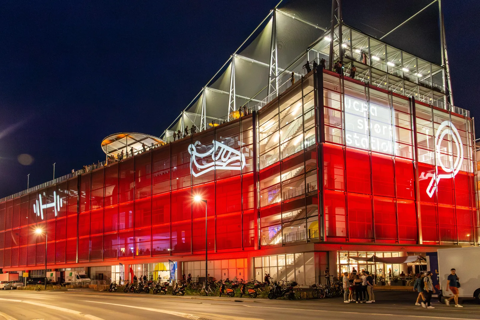 Vue nocturne de l'UCPA Sport Station à Bordeaux, un équipement sportif moderne aux façades rouges et vitrées illuminées, avec des visiteurs devant l'entrée