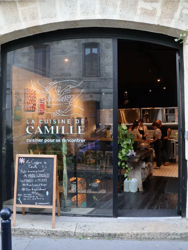 Devanture du restaurant La Cuisine de Camille à Bordeaux, avec de grandes vitrines montrant l'intérieur éclairé où les clients dînent, et un menu sur ardoise à l'extérieur sur la rue.