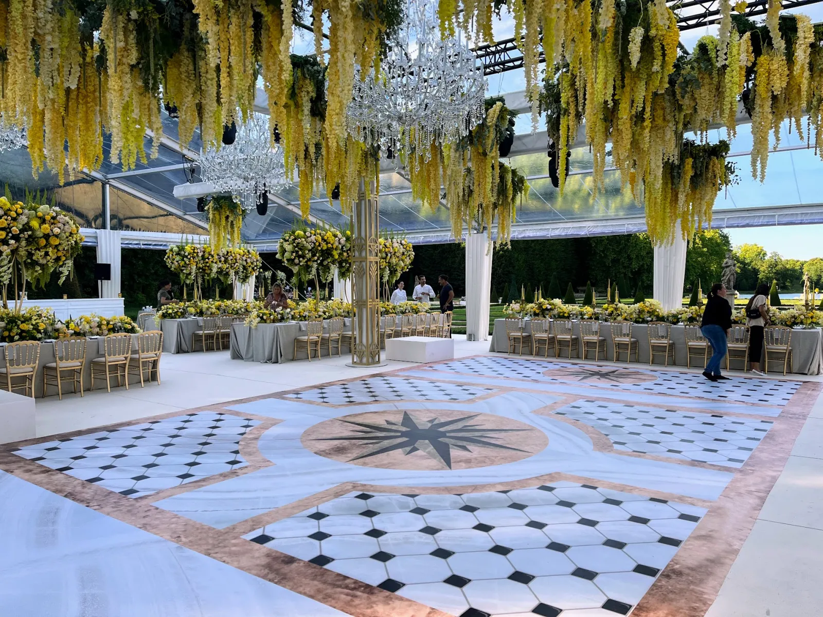 Lieu de réception de mariage élégant à Bordeaux avec un sol aux motifs géométriques, des fleurs de glycine jaune suspendues, un lustre en cristal, des tables rondes avec des nappes grises et des haies taillées sous un pavillon à toit transparent.