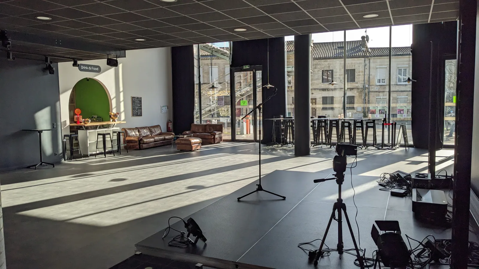 Espace événementiel contemporain de L'Estrade à Bordeaux équipé de matériel d'éclairage professionnel, zone de sièges en cuir marron, mur d'accent vert avec enseigne 'Drink & Food', et fenêtres panoramiques sur les immeubles haussmanniens