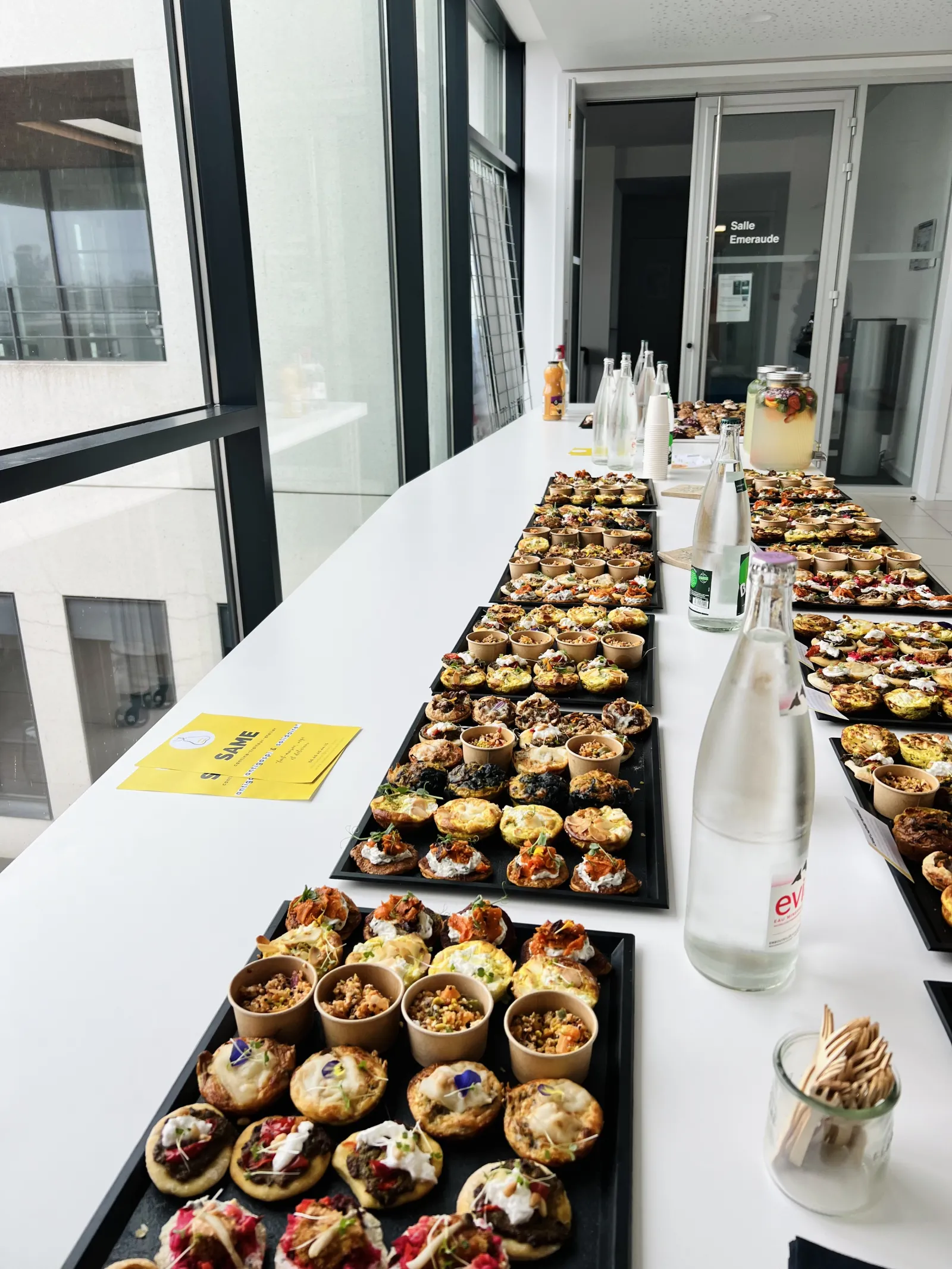 Une longue table blanche présentant plusieurs plateaux noirs garnis de canapés gourmet aux garnitures variées, accompagnés de boissons et condiments dans un espace moderne avec baies vitrées.