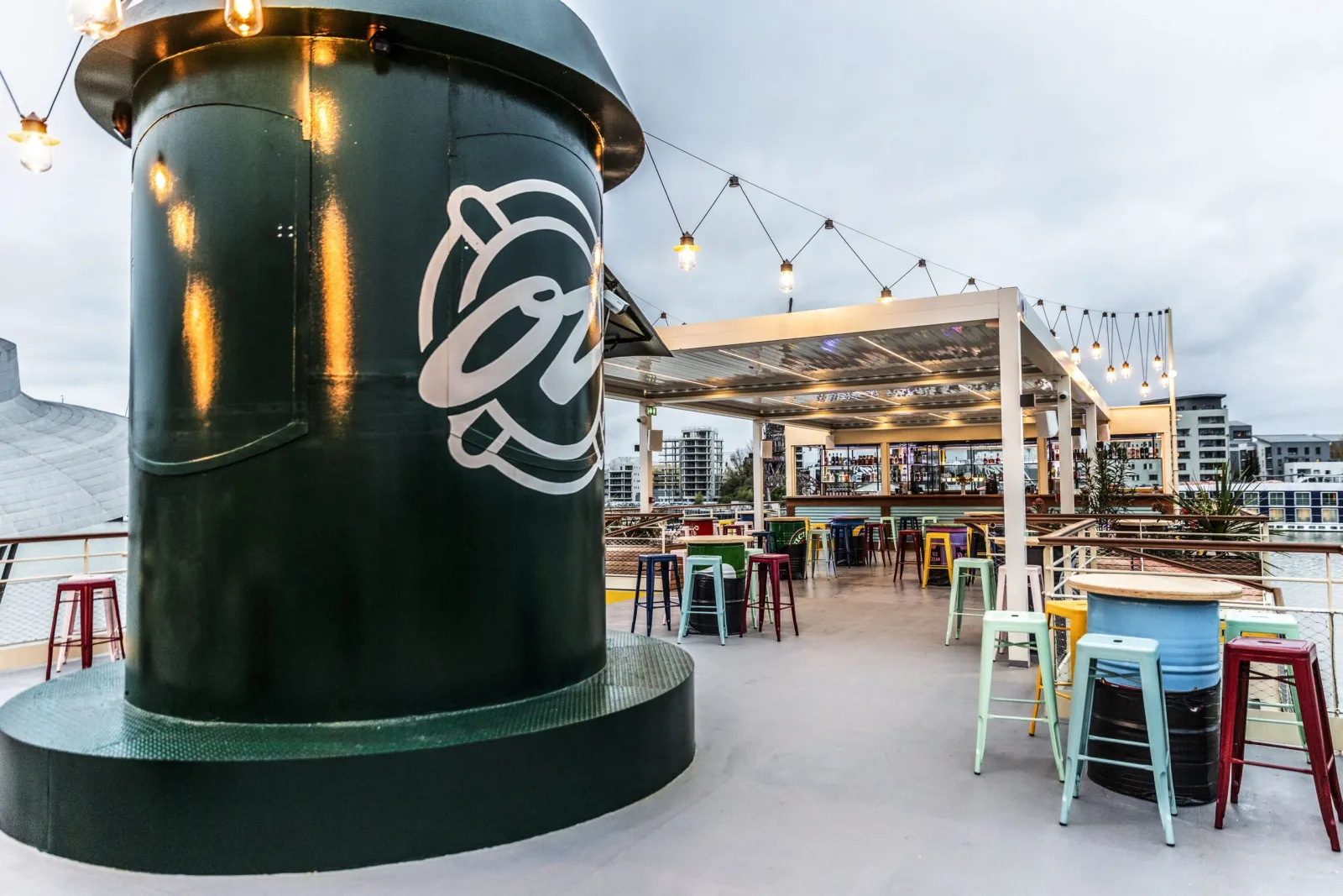 Terrasse extérieure en bord de l'eau à OZ Boat à Bordeaux, avec une structure cylindrique verte ornée du logo, des tabourets de bar colorés, une pergola moderne en bois avec luminaires suspendus, et immeubles urbains en arrière-plan.