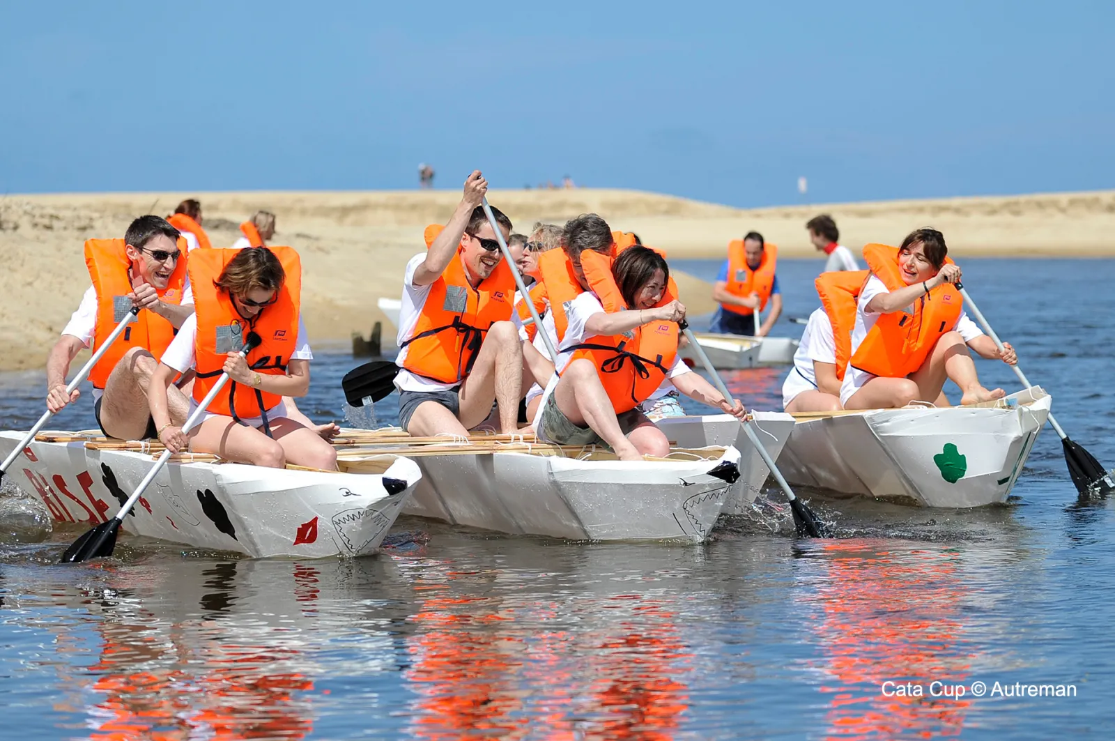 Groupe de personnes portant des gilets de sauvetage orange ramant dans de petits bateaux blancs avec des pagaies en eau côtière peu profonde, avec plage de sable et ciel bleu en arrière-plan.