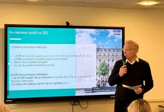 Un présentateur en pull noir tient un microphone devant un écran affichant une présentation sur les indicateurs positifs de l'hôtellerie à Bordeaux Métropole en 2023, incluant les taux d'occupation, les chiffres de revenus et les statistiques touristiques