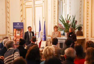 Événement formel dans un salon orné de Bordeaux avec des intervenants à un pupitre aux côtés des drapeaux français et européen, s'adressant à un public dans une salle élégante aux décors dorés et arrangement floral.