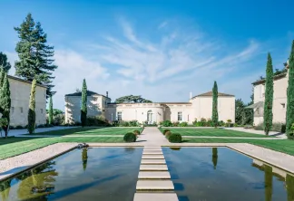 Domaine élégant de style néoclassique à Bordeaux avec des bâtiments symétriques de couleur crème, des cyprès, une pelouse centrale et un bassin de réflexion traversé par un chemin de pierre.