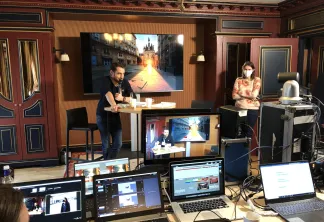 Configuration d'un événement virtuel à Bordeaux avec deux participants à un bureau, plusieurs écrans affichant un décor urbain, et équipements de production incluant ordinateurs portables et moniteurs pour une conférence à distance.